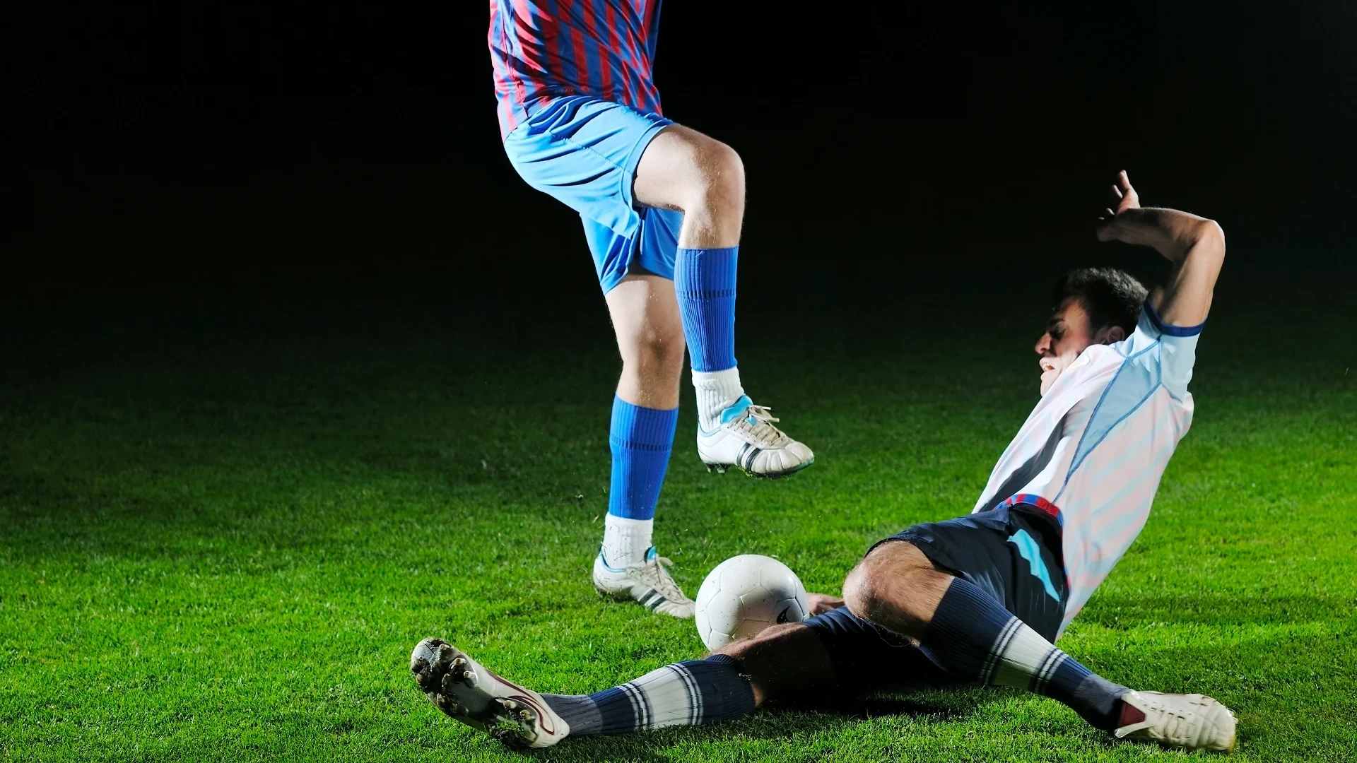 Two footballs are about to collide badly, trying to fight for the ball. The player on the bottom is just about to get studs printed onto his arm.